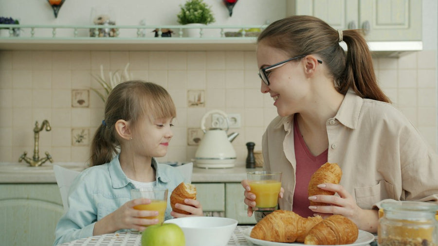 Mamá e hija desayunan juntas en la cocina, con jugo y croissants