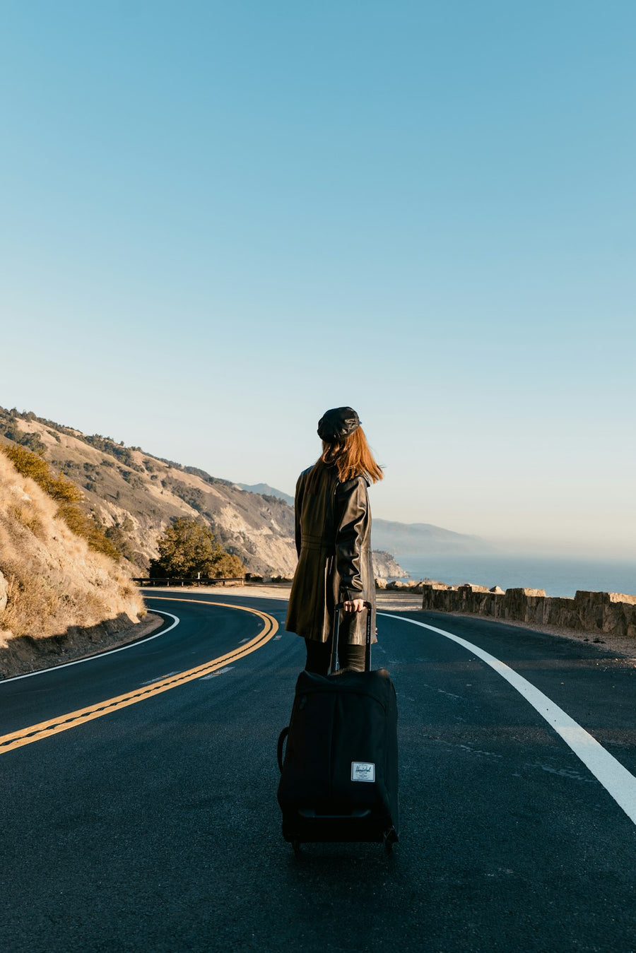 Mujer con chaqueta café caminando por una carretera junto al mar